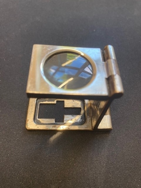 Sammenleggbar lupe i etui av lær. Lupen foldes opp slik at områder på 5mm og 10mm og en tomme vises i synsfeltet. En kan så telle hvor mange tråder er på dette området, for å kvalitetssikre tekstilet. På stativets utside er logo fra Haldens gravert i overflaten.