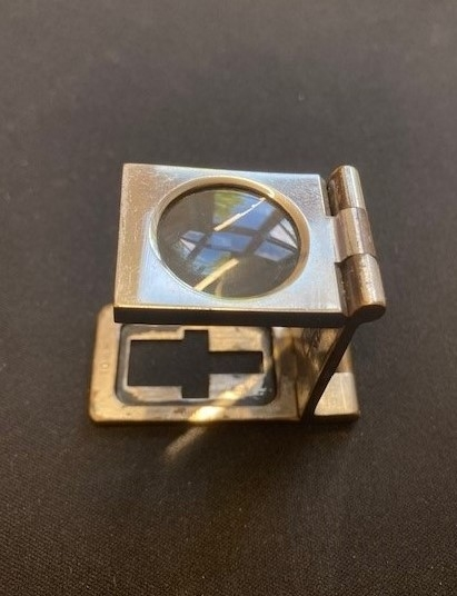 Sammenleggbar lupe i etui av lær. Lupen foldes opp slik at områder på 5mm og 10mm og en tomme vises i synsfeltet. En kan så telle hvor mange tråder er på dette området, for å kvalitetssikre tekstilet. På stativets utside er logo fra Haldens gravert i overflaten.