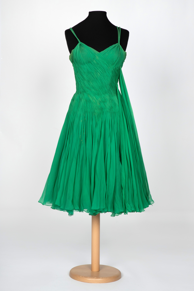 Kort, ermeløs cocktailkjole av grønn silkechiffon. Kjolen er utringet og med tettsittende liv med 9 spiler, utbygget over bysten og med lang glidelås i venstre side. Vidt, struttende skjørt og innsvinget liv. Kjolen består av flere lag stoff: ytterst et lag tynn chiffon, plissert og diagonalt drapert og lett stukket ned inntil hoftelengde. Herfra faller stoffet fritt i et vidt skjørt med et lag av tynn, ukreppet chiffon under. Underst en underkjole av grønn taft. To rouleauer er stukket mellom stofflengdene over bysten og holder kjolen oppe over skuldrene. To løse, plisserte panel på kjolens venstre side.