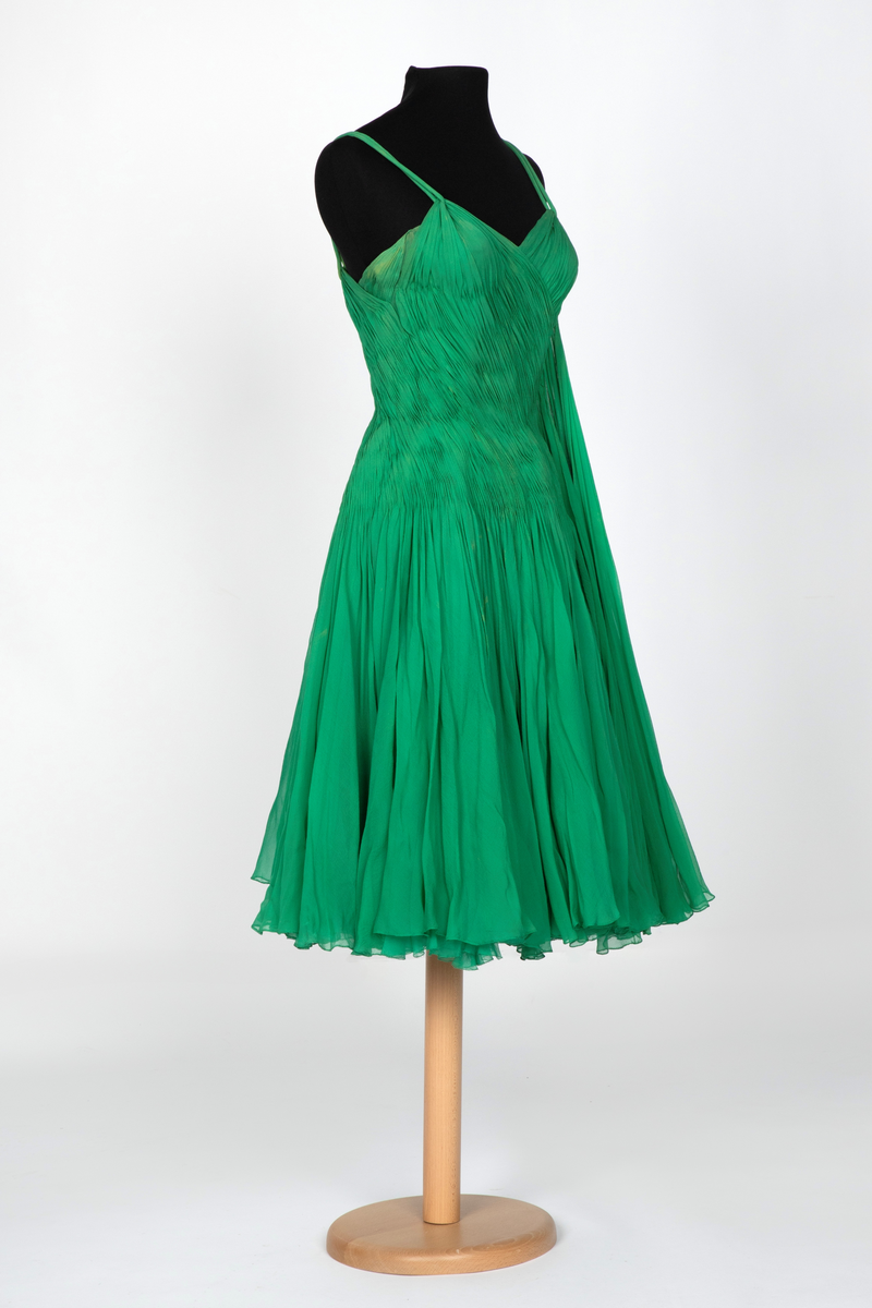 Kort, ermeløs cocktailkjole av grønn silkechiffon. Kjolen er utringet og med tettsittende liv med 9 spiler, utbygget over bysten og med lang glidelås i venstre side. Vidt, struttende skjørt og innsvinget liv. Kjolen består av flere lag stoff: ytterst et lag tynn chiffon, plissert og diagonalt drapert og lett stukket ned inntil hoftelengde. Herfra faller stoffet fritt i et vidt skjørt med et lag av tynn, ukreppet chiffon under. Underst en underkjole av grønn taft. To rouleauer er stukket mellom stofflengdene over bysten og holder kjolen oppe over skuldrene. To løse, plisserte panel på kjolens venstre side.