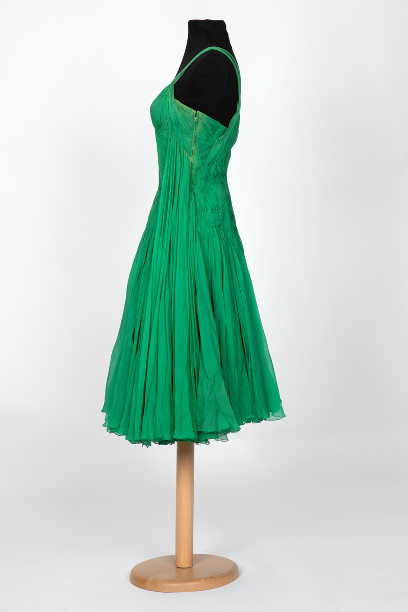 Kort, ermeløs cocktailkjole av grønn silkechiffon. Kjolen er utringet og med tettsittende liv med 9 spiler, utbygget over bysten og med lang glidelås i venstre side. Vidt, struttende skjørt og innsvinget liv. Kjolen består av flere lag stoff: ytterst et lag tynn chiffon, plissert og diagonalt drapert og lett stukket ned inntil hoftelengde. Herfra faller stoffet fritt i et vidt skjørt med et lag av tynn, ukreppet chiffon under. Underst en underkjole av grønn taft. To rouleauer er stukket mellom stofflengdene over bysten og holder kjolen oppe over skuldrene. To løse, plisserte panel på kjolens venstre side.
