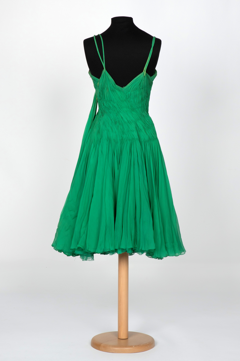 Kort, ermeløs cocktailkjole av grønn silkechiffon. Kjolen er utringet og med tettsittende liv med 9 spiler, utbygget over bysten og med lang glidelås i venstre side. Vidt, struttende skjørt og innsvinget liv. Kjolen består av flere lag stoff: ytterst et lag tynn chiffon, plissert og diagonalt drapert og lett stukket ned inntil hoftelengde. Herfra faller stoffet fritt i et vidt skjørt med et lag av tynn, ukreppet chiffon under. Underst en underkjole av grønn taft. To rouleauer er stukket mellom stofflengdene over bysten og holder kjolen oppe over skuldrene. To løse, plisserte panel på kjolens venstre side.