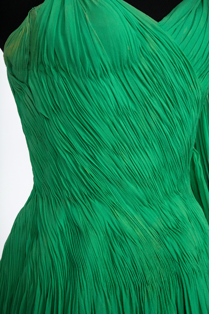 Kort, ermeløs cocktailkjole av grønn silkechiffon. Kjolen er utringet og med tettsittende liv med 9 spiler, utbygget over bysten og med lang glidelås i venstre side. Vidt, struttende skjørt og innsvinget liv. Kjolen består av flere lag stoff: ytterst et lag tynn chiffon, plissert og diagonalt drapert og lett stukket ned inntil hoftelengde. Herfra faller stoffet fritt i et vidt skjørt med et lag av tynn, ukreppet chiffon under. Underst en underkjole av grønn taft. To rouleauer er stukket mellom stofflengdene over bysten og holder kjolen oppe over skuldrene. To løse, plisserte panel på kjolens venstre side.