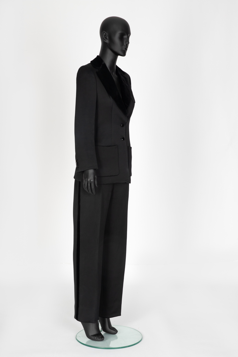 Smoking i svart silke- og ullsateng. Jakken har speilfløyelsslag og galloner. Enkeltspent med to knapper og tre lommer. Buksen har rette ben. Den er sydd ut i livet og linningen skjøtet på midt bak. Har hatt legg eller innsnitt foran. Tynn stripe i fløyel på siden av hvert bein.