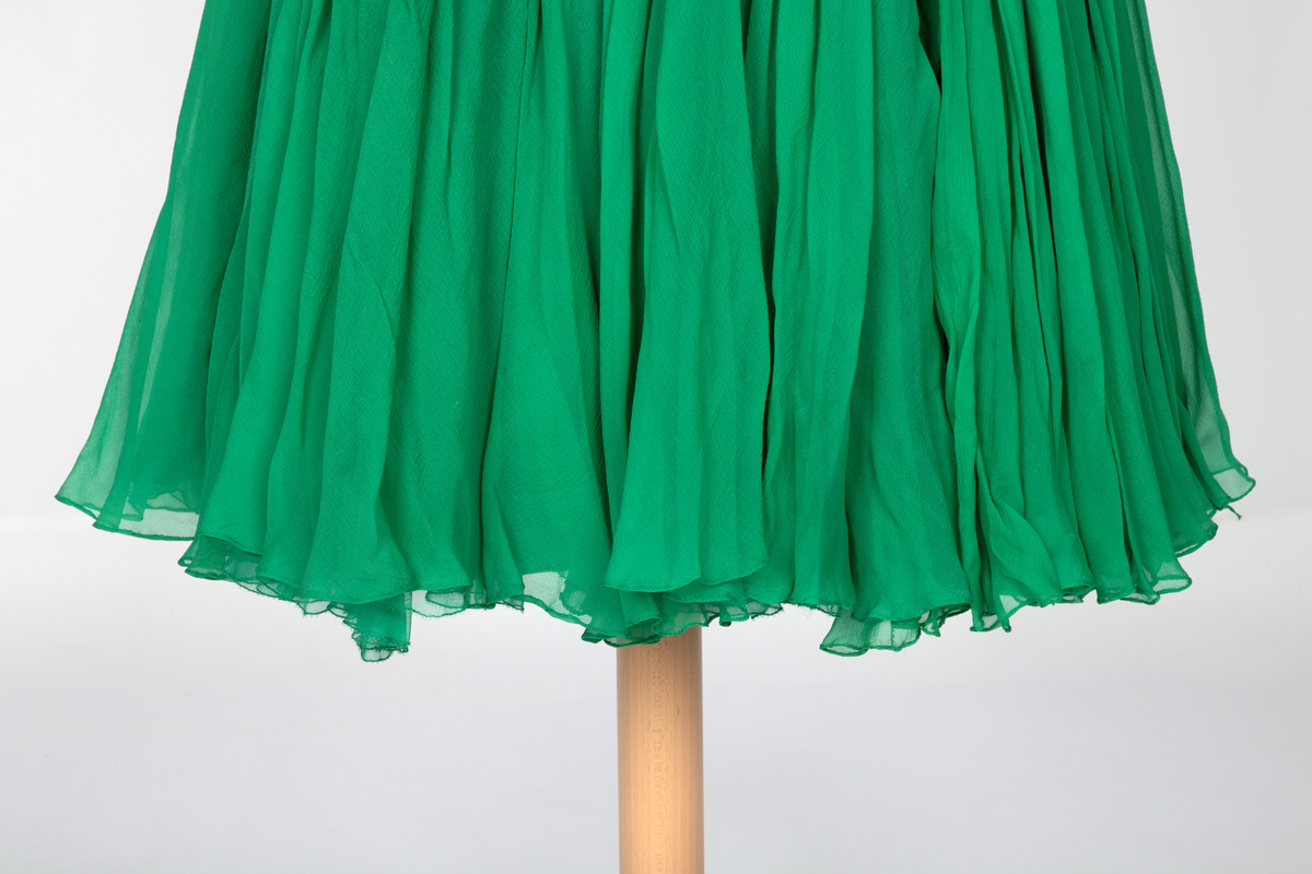 Kort, ermeløs cocktailkjole av grønn silkechiffon. Kjolen er utringet og med tettsittende liv med 9 spiler, utbygget over bysten og med lang glidelås i venstre side. Vidt, struttende skjørt og innsvinget liv. Kjolen består av flere lag stoff: ytterst et lag tynn chiffon, plissert og diagonalt drapert og lett stukket ned inntil hoftelengde. Herfra faller stoffet fritt i et vidt skjørt med et lag av tynn, ukreppet chiffon under. Underst en underkjole av grønn taft. To rouleauer er stukket mellom stofflengdene over bysten og holder kjolen oppe over skuldrene. To løse, plisserte panel på kjolens venstre side.