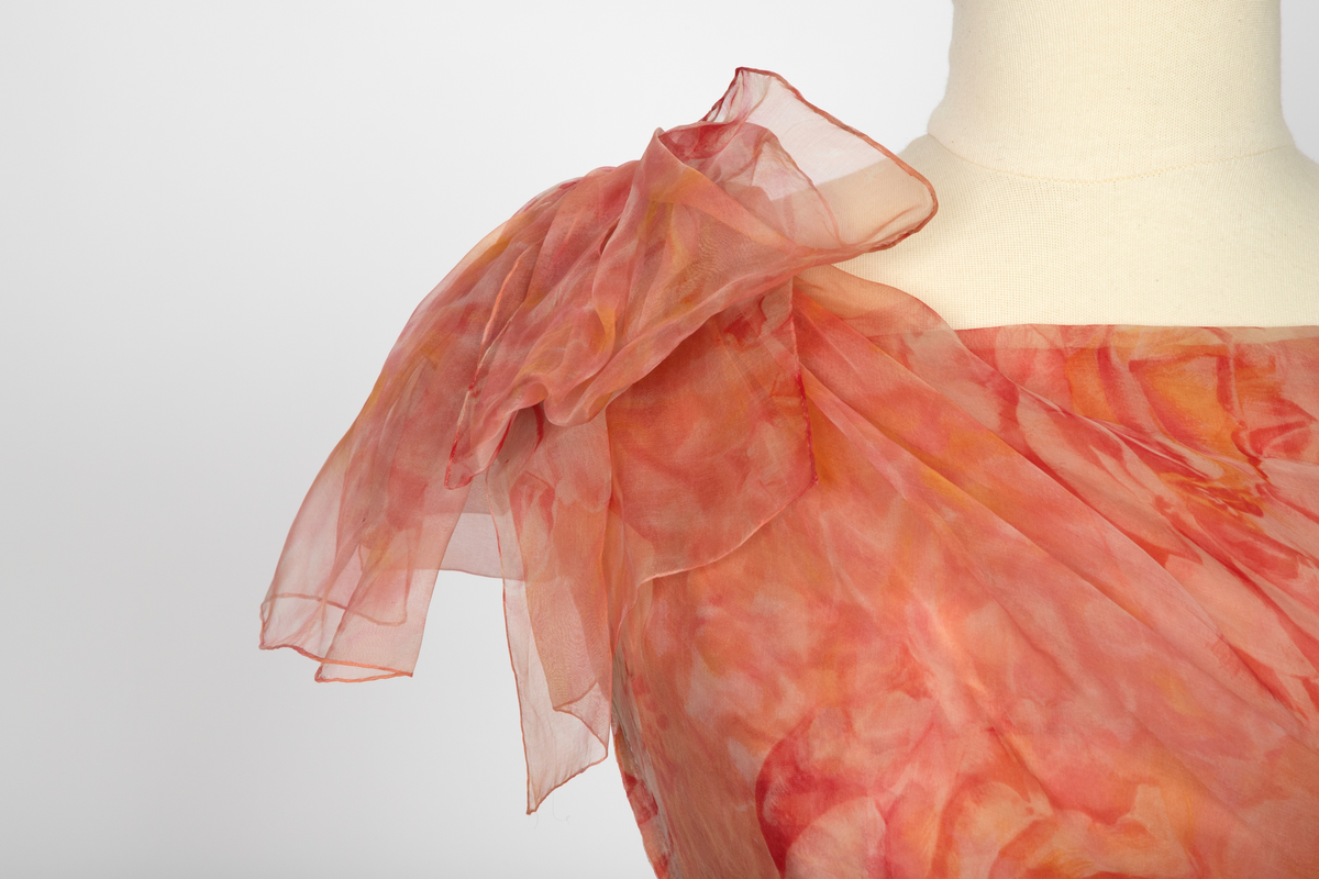 Kort, ermeløs cocktailkjole av blomstret chiffon i lyserøde og gule nyanser. Kjolen har båtformet utringning og lav livlinje. Skjørtet består av tre lag blomstret chiffon, småfoldet og med stor vidde i de to ytterste lag, det tredje noe trangere og med et tverrbånd av stivt nettstoff på nedre del. Under dette et lag av umønstret chiffon. Underst en hvit silkeunderkjole med hvit chiffonkant nederst. Livet har et ytre chiffonlag, diagonalt drapert og samlet under en stor sløyfe på høyre skulder. Kjolen har glidelås i ryggen og er hektet og knappet i høyre side.