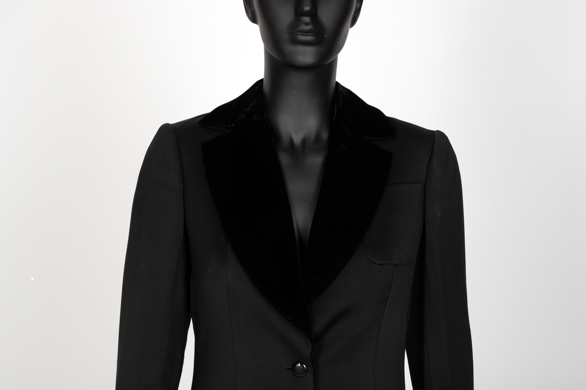 Smoking i svart silke- og ullsateng. Jakken har speilfløyelsslag og galloner. Enkeltspent med to knapper og tre lommer. Buksen har rette ben. Den er sydd ut i livet og linningen skjøtet på midt bak. Har hatt legg eller innsnitt foran. Tynn stripe i fløyel på siden av hvert bein.