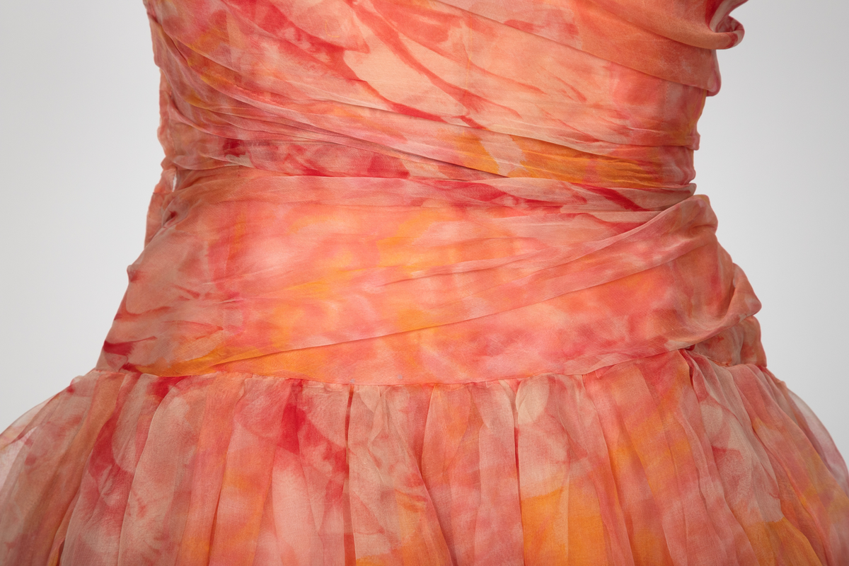 Kort, ermeløs cocktailkjole av blomstret chiffon i lyserøde og gule nyanser. Kjolen har båtformet utringning og lav livlinje. Skjørtet består av tre lag blomstret chiffon, småfoldet og med stor vidde i de to ytterste lag, det tredje noe trangere og med et tverrbånd av stivt nettstoff på nedre del. Under dette et lag av umønstret chiffon. Underst en hvit silkeunderkjole med hvit chiffonkant nederst. Livet har et ytre chiffonlag, diagonalt drapert og samlet under en stor sløyfe på høyre skulder. Kjolen har glidelås i ryggen og er hektet og knappet i høyre side.