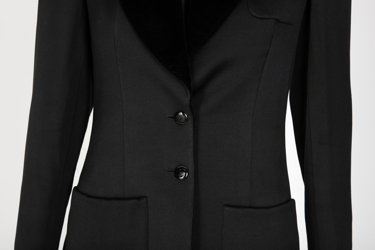 Smoking i svart silke- og ullsateng. Jakken har speilfløyelsslag og galloner. Enkeltspent med to knapper og tre lommer. Buksen har rette ben. Den er sydd ut i livet og linningen skjøtet på midt bak. Har hatt legg eller innsnitt foran. Tynn stripe i fløyel på siden av hvert bein.