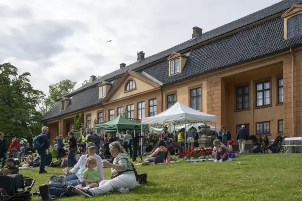 Besøkende nyter mat og aktiviteter foran salgsboder og hovedbygningen på Bogstad Gård under Sankthansfeiring.