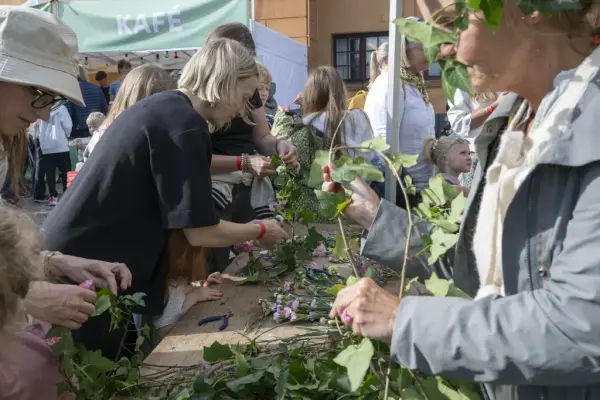 Barn og voksne lager blomsterkranser med eføy og markblomster under Sankthansfeiring på Bogstad Gård.