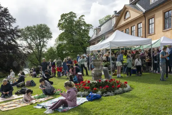 Folk samles rundt boder og på pikniktepper foran Bogstad Gård under Sankthansfeiring med blomsterbed i forgrunnen.