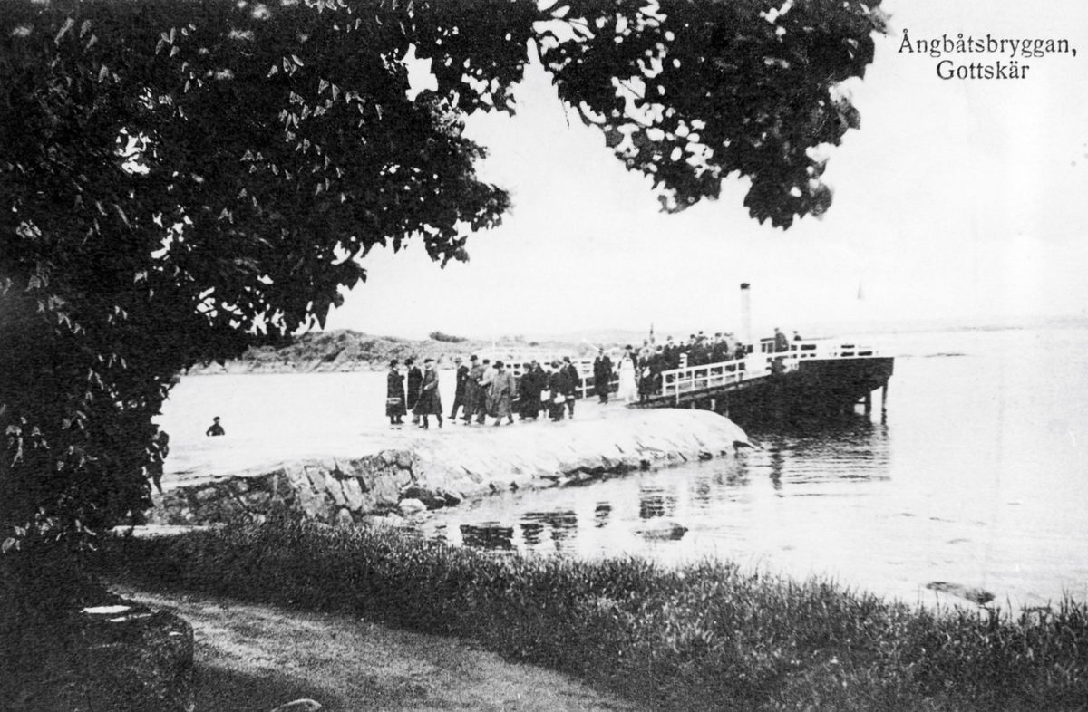 Vykort, "Ångbåtsbryggan, Gottskär." En rad passagerare går på piren från ångbåten in mot land. Gottskär var en livlig badort och båttrafiken dit utgick från Kungsbacka.
