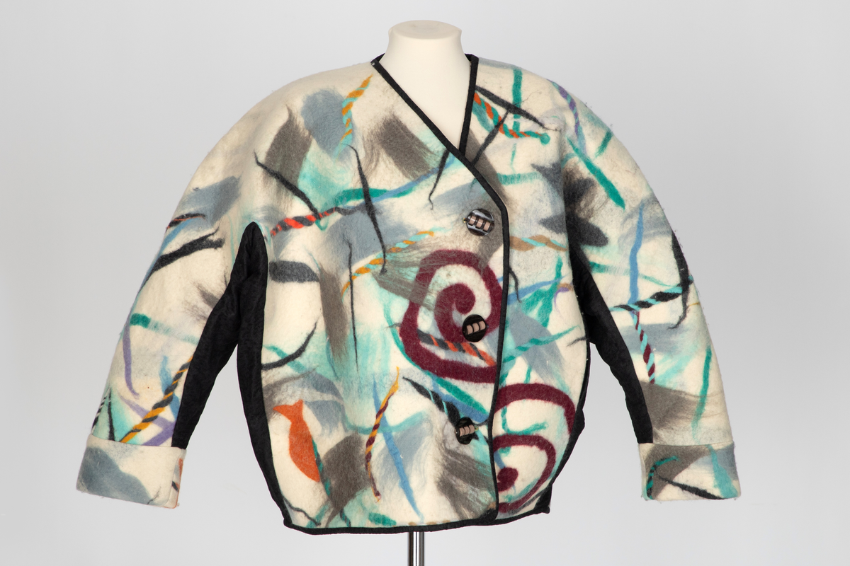Jakke av filtet (tovet) ull. Ullstoffet har innlagte tråder i mange farger. Kimonofasong med innfelte stykker av blondestoff i sidene og under ermene. Halsen har asymmetrisk lukning mot venstre og jakken knappes foran med tre store plastknapper i passepoilerte knapphull. Forkanten på jakken har svarte kanter av blondestoffet. Jakken har rutete bomullsfôr.