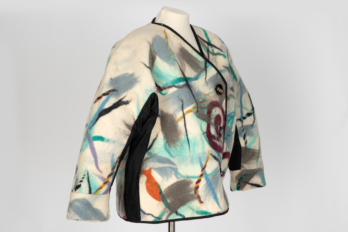 Jakke av filtet (tovet) ull. Ullstoffet har innlagte tråder i mange farger. Kimonofasong med innfelte stykker av blondestoff i sidene og under ermene. Halsen har asymmetrisk lukning mot venstre og jakken knappes foran med tre store plastknapper i passepoilerte knapphull. Forkanten på jakken har svarte kanter av blondestoffet. Jakken har rutete bomullsfôr.