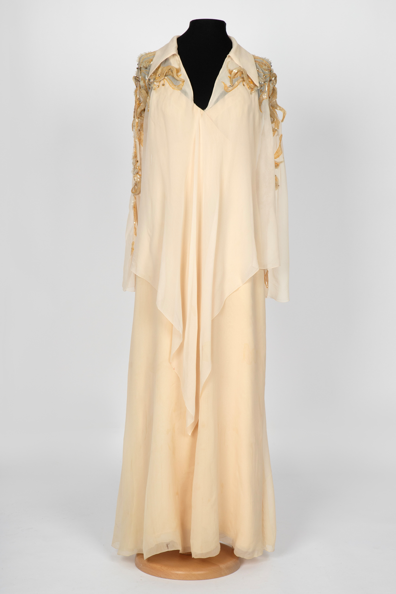 Aftenkjole med poncho. Kjolen er i elfenbensfarget silkechiffon med utskrådt, ankelsidt skjørt, v-ringning og smale skulderstropper. Glidelås i ryggen. Ponchoen har en kort splitt i halsen og spiss krage. Skuldre og nedover ermene er dekorert med gullperler, blanke paljetter og stener formet til bladverk, noen opphøyd.