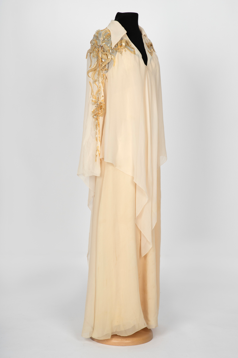 Aftenkjole med poncho. Kjolen er i elfenbensfarget silkechiffon med utskrådt, ankelsidt skjørt, v-ringning og smale skulderstropper. Glidelås i ryggen. Ponchoen har en kort splitt i halsen og spiss krage. Skuldre og nedover ermene er dekorert med gullperler, blanke paljetter og stener formet til bladverk, noen opphøyd.