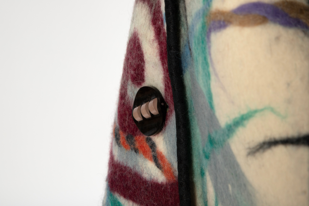 Jakke av filtet (tovet) ull. Ullstoffet har innlagte tråder i mange farger. Kimonofasong med innfelte stykker av blondestoff i sidene og under ermene. Halsen har asymmetrisk lukning mot venstre og jakken knappes foran med tre store plastknapper i passepoilerte knapphull. Forkanten på jakken har svarte kanter av blondestoffet. Jakken har rutete bomullsfôr.