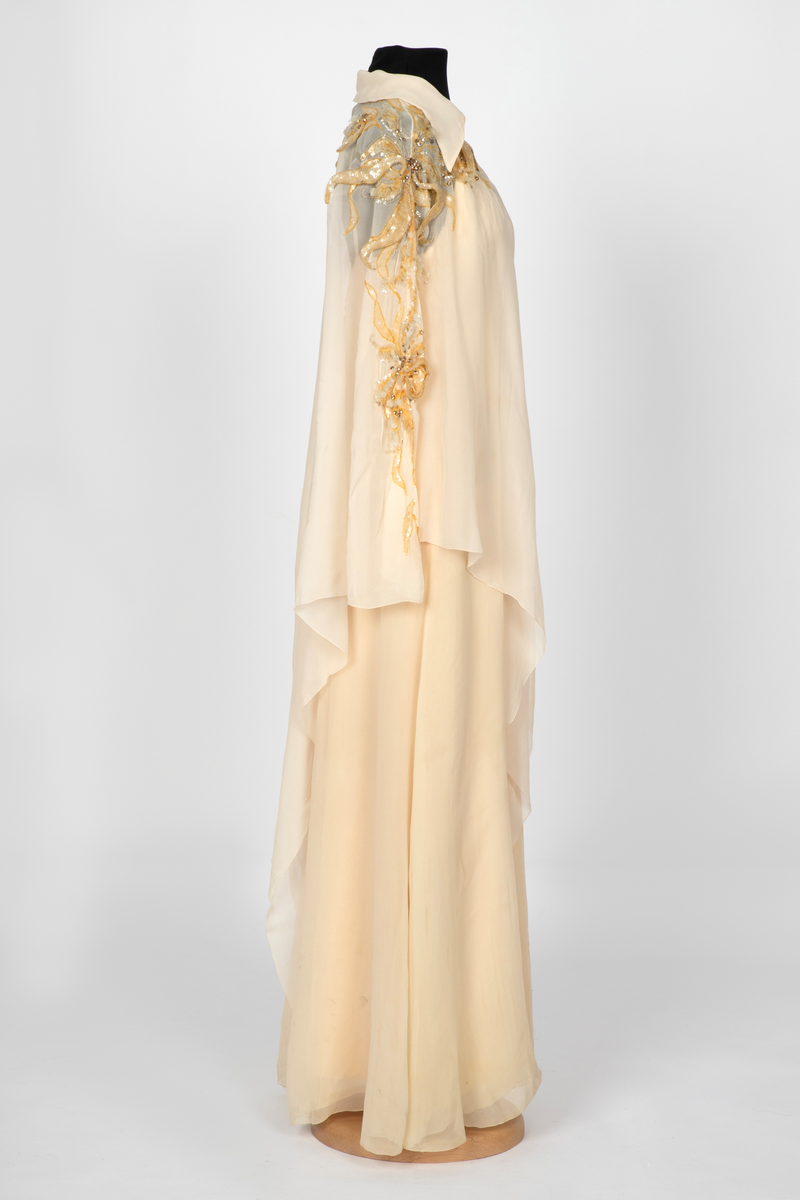 Aftenkjole med poncho. Kjolen er i elfenbensfarget silkechiffon med utskrådt, ankelsidt skjørt, v-ringning og smale skulderstropper. Glidelås i ryggen. Ponchoen har en kort splitt i halsen og spiss krage. Skuldre og nedover ermene er dekorert med gullperler, blanke paljetter og stener formet til bladverk, noen opphøyd.
