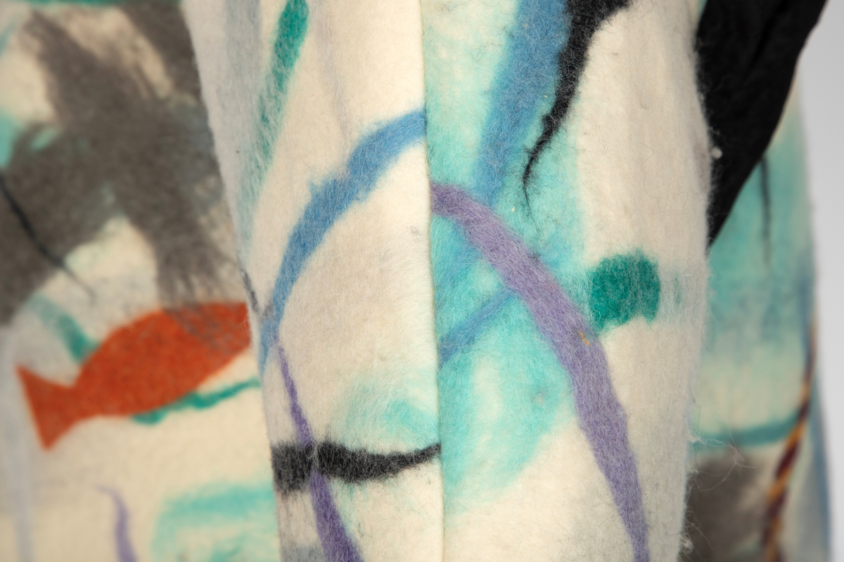 Jakke av filtet (tovet) ull. Ullstoffet har innlagte tråder i mange farger. Kimonofasong med innfelte stykker av blondestoff i sidene og under ermene. Halsen har asymmetrisk lukning mot venstre og jakken knappes foran med tre store plastknapper i passepoilerte knapphull. Forkanten på jakken har svarte kanter av blondestoffet. Jakken har rutete bomullsfôr.