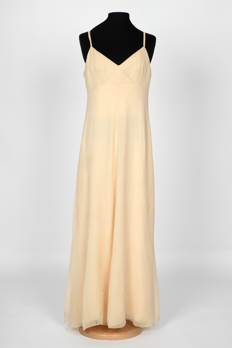 Aftenkjole i elfenbensfarget silkechiffon med utskrådt, ankelsidt skjørt, v-ringning og smale skulderstropper. Glidelås i ryggen.