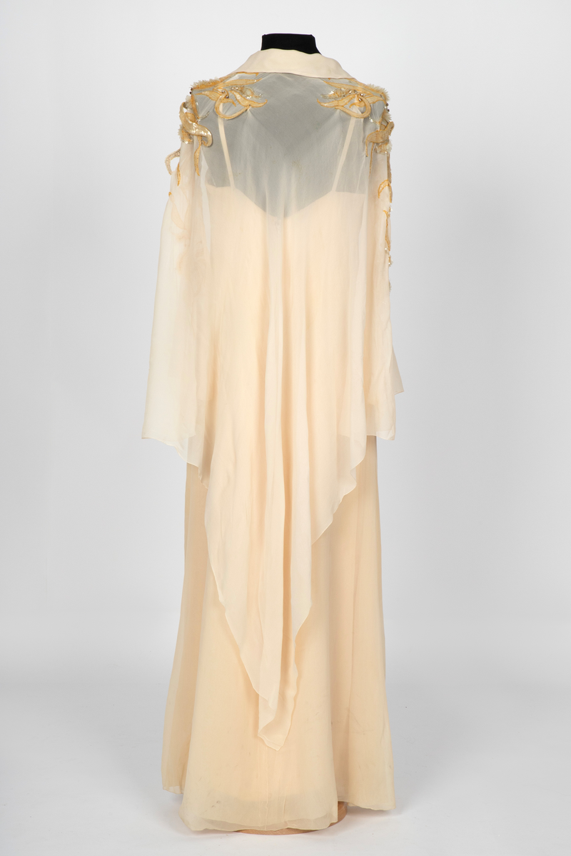Aftenkjole med poncho. Kjolen er i elfenbensfarget silkechiffon med utskrådt, ankelsidt skjørt, v-ringning og smale skulderstropper. Glidelås i ryggen. Ponchoen har en kort splitt i halsen og spiss krage. Skuldre og nedover ermene er dekorert med gullperler, blanke paljetter og stener formet til bladverk, noen opphøyd.