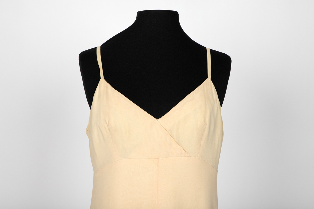 Aftenkjole i elfenbensfarget silkechiffon med utskrådt, ankelsidt skjørt, v-ringning og smale skulderstropper. Glidelås i ryggen.