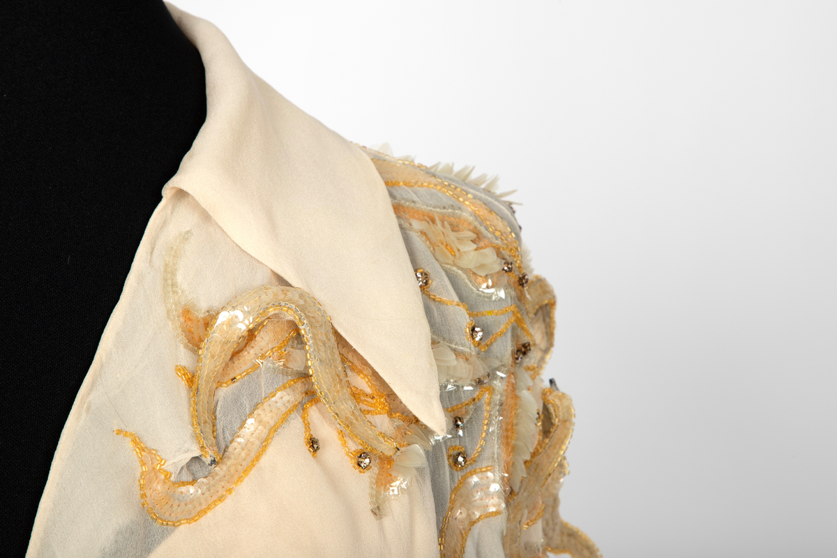 Aftenkjole med poncho. Kjolen er i elfenbensfarget silkechiffon med utskrådt, ankelsidt skjørt, v-ringning og smale skulderstropper. Glidelås i ryggen. Ponchoen har en kort splitt i halsen og spiss krage. Skuldre og nedover ermene er dekorert med gullperler, blanke paljetter og stener formet til bladverk, noen opphøyd.