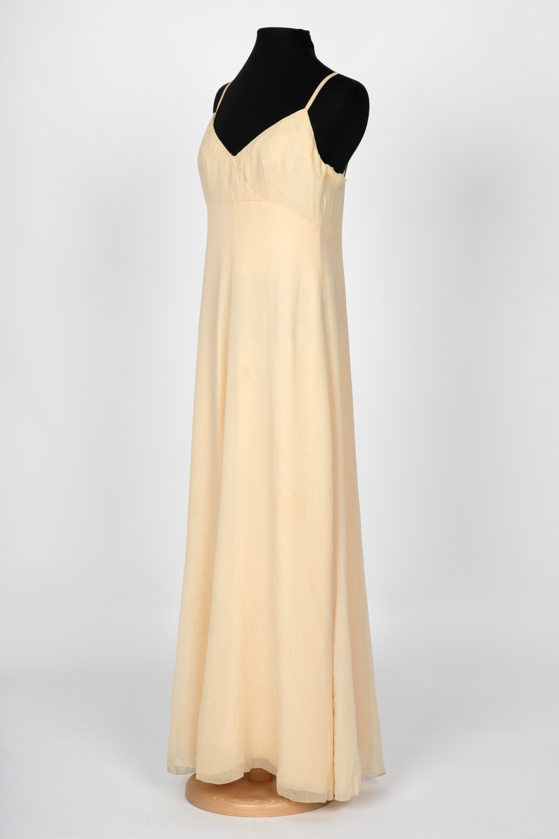 Aftenkjole i elfenbensfarget silkechiffon med utskrådt, ankelsidt skjørt, v-ringning og smale skulderstropper. Glidelås i ryggen.