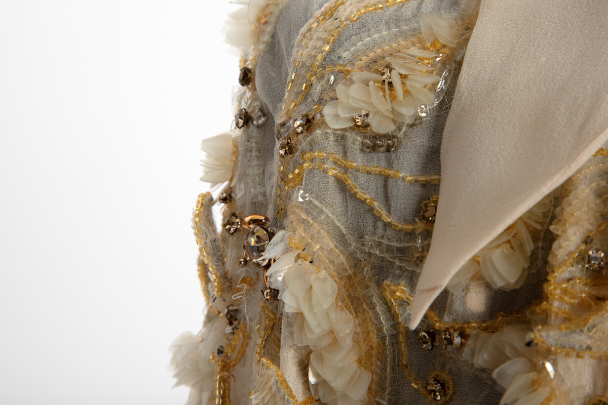 Aftenkjole med poncho. Kjolen er i elfenbensfarget silkechiffon med utskrådt, ankelsidt skjørt, v-ringning og smale skulderstropper. Glidelås i ryggen. Ponchoen har en kort splitt i halsen og spiss krage. Skuldre og nedover ermene er dekorert med gullperler, blanke paljetter og stener formet til bladverk, noen opphøyd.