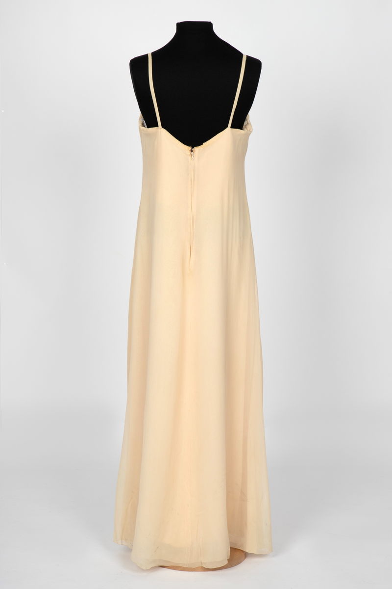 Aftenkjole i elfenbensfarget silkechiffon med utskrådt, ankelsidt skjørt, v-ringning og smale skulderstropper. Glidelås i ryggen.