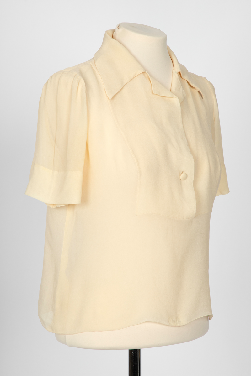 Bluse i elfenbensfarget silkechiffon med firkantet skjæring midt foran. Enkel knepping med en silketrukket knapp. Skjorteblusekrage med slag. To knapper er festet midt foran og midt bak i den nedre kanten.