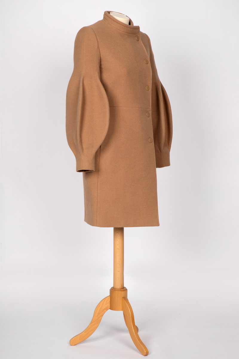 3/4 lang jakke eller kort kåpe i beige ullstoff med overdimensjonerte ballongermer. Jakken har innsvinget midje, to stikklommer i sidene og asymmetrisk lukning med fem knapper.