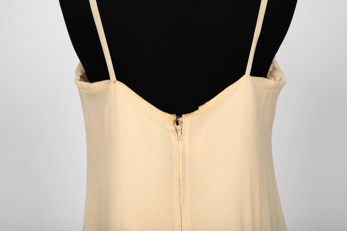 Aftenkjole i elfenbensfarget silkechiffon med utskrådt, ankelsidt skjørt, v-ringning og smale skulderstropper. Glidelås i ryggen.