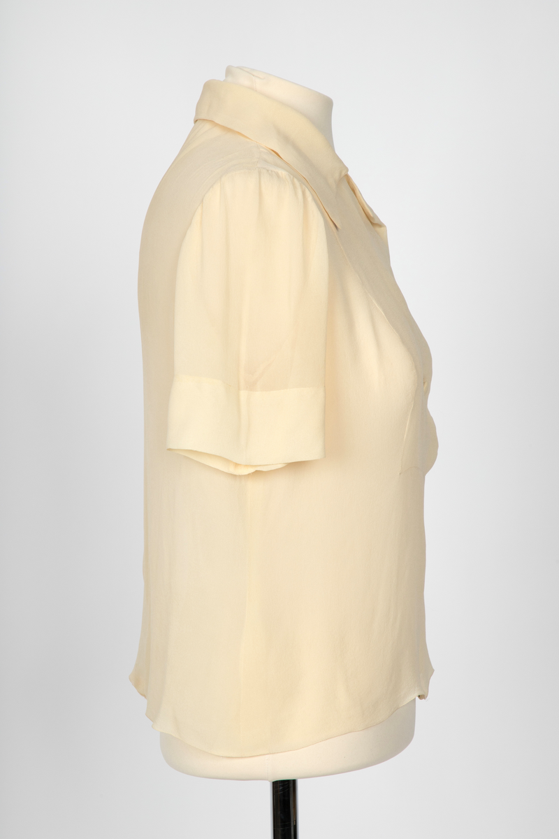 Bluse i elfenbensfarget silkechiffon med firkantet skjæring midt foran. Enkel knepping med en silketrukket knapp. Skjorteblusekrage med slag. To knapper er festet midt foran og midt bak i den nedre kanten.