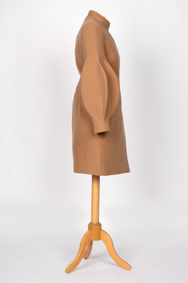 3/4 lang jakke eller kort kåpe i beige ullstoff med overdimensjonerte ballongermer. Jakken har innsvinget midje, to stikklommer i sidene og asymmetrisk lukning med fem knapper.