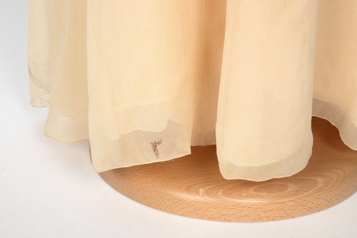 Aftenkjole i elfenbensfarget silkechiffon med utskrådt, ankelsidt skjørt, v-ringning og smale skulderstropper. Glidelås i ryggen.