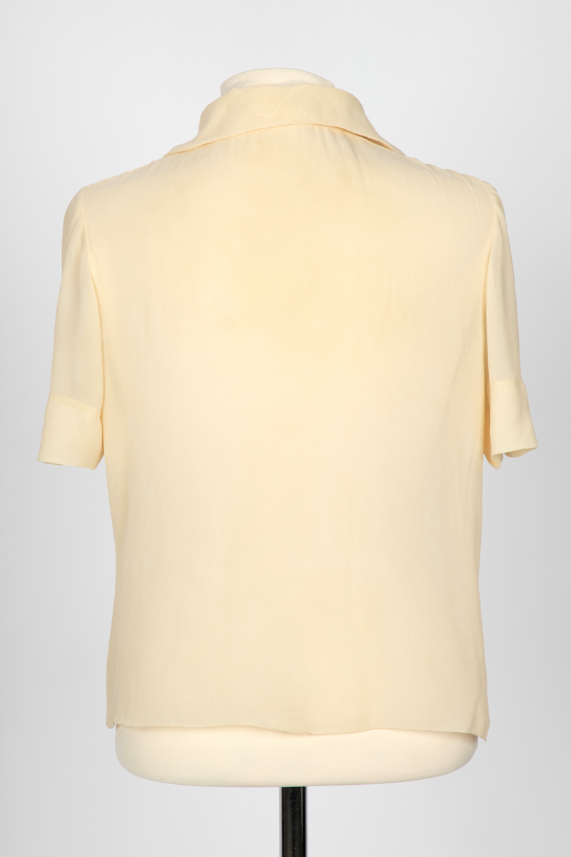 Bluse i elfenbensfarget silkechiffon med firkantet skjæring midt foran. Enkel knepping med en silketrukket knapp. Skjorteblusekrage med slag. To knapper er festet midt foran og midt bak i den nedre kanten.