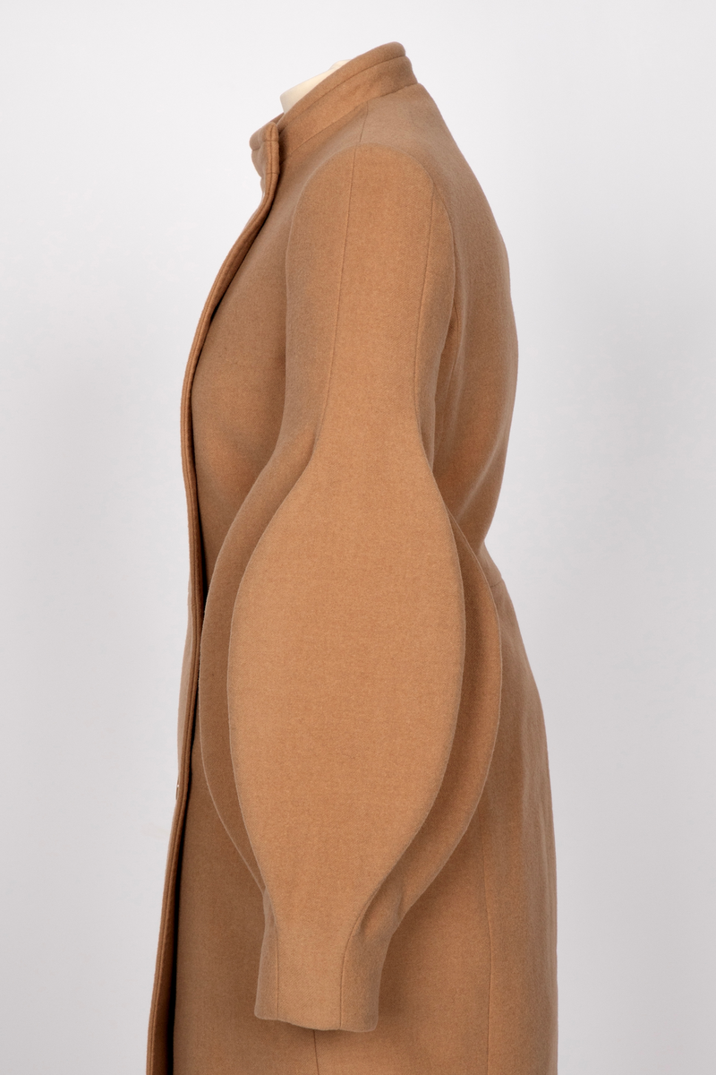 3/4 lang jakke eller kort kåpe i beige ullstoff med overdimensjonerte ballongermer. Jakken har innsvinget midje, to stikklommer i sidene og asymmetrisk lukning med fem knapper.