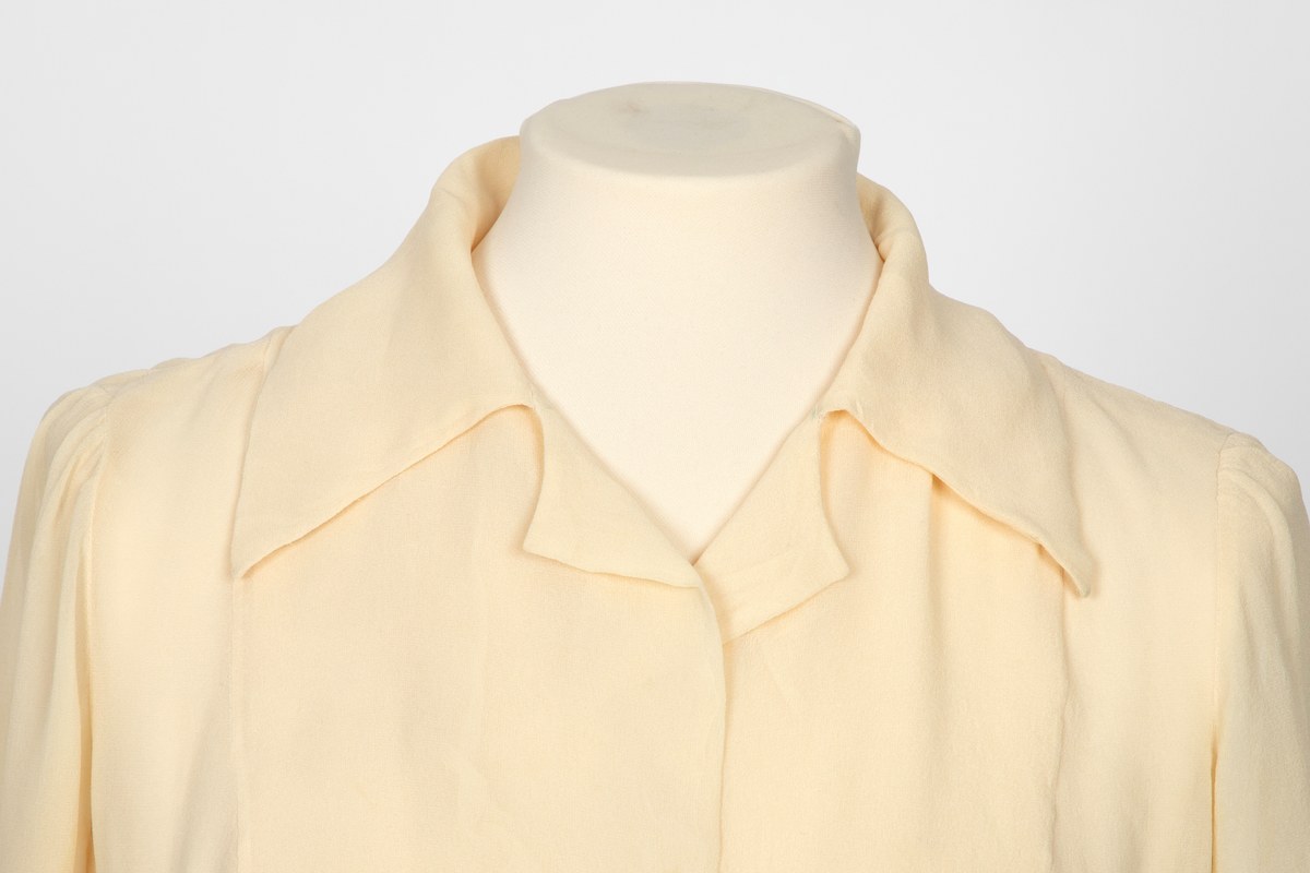 Bluse i elfenbensfarget silkechiffon med firkantet skjæring midt foran. Enkel knepping med en silketrukket knapp. Skjorteblusekrage med slag. To knapper er festet midt foran og midt bak i den nedre kanten.