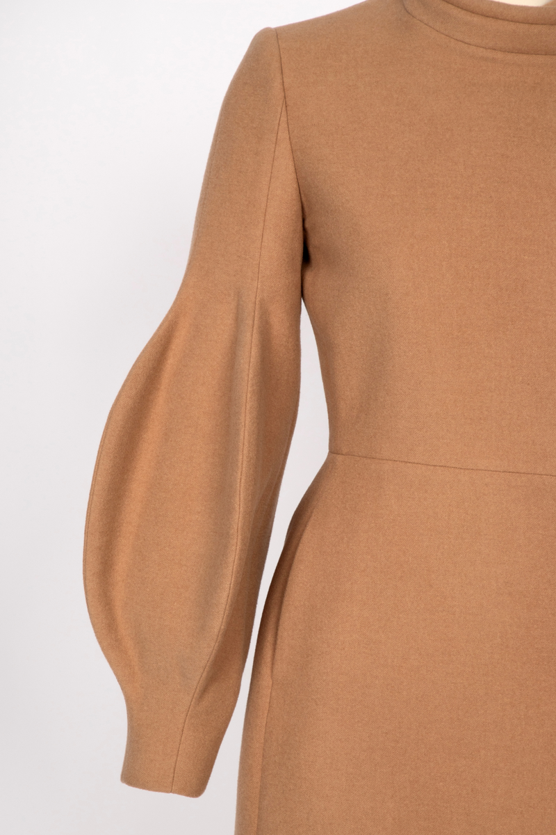 3/4 lang jakke eller kort kåpe i beige ullstoff med overdimensjonerte ballongermer. Jakken har innsvinget midje, to stikklommer i sidene og asymmetrisk lukning med fem knapper.