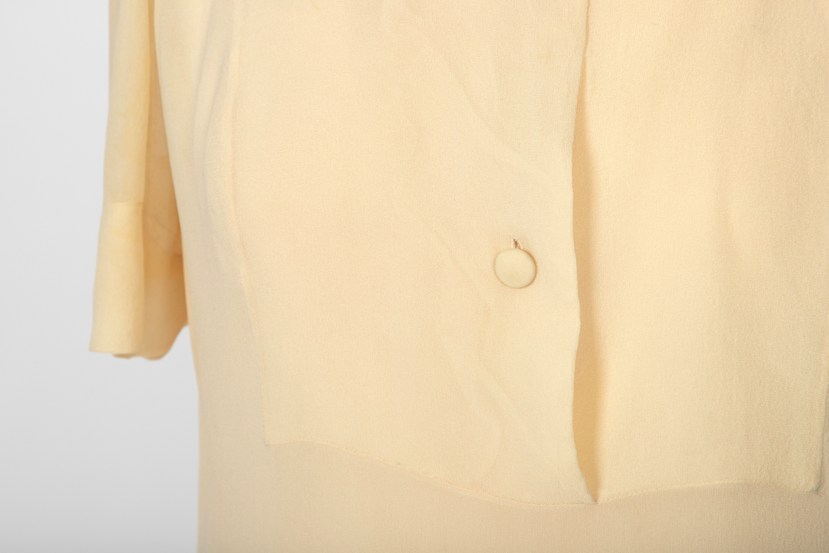 Bluse i elfenbensfarget silkechiffon med firkantet skjæring midt foran. Enkel knepping med en silketrukket knapp. Skjorteblusekrage med slag. To knapper er festet midt foran og midt bak i den nedre kanten.