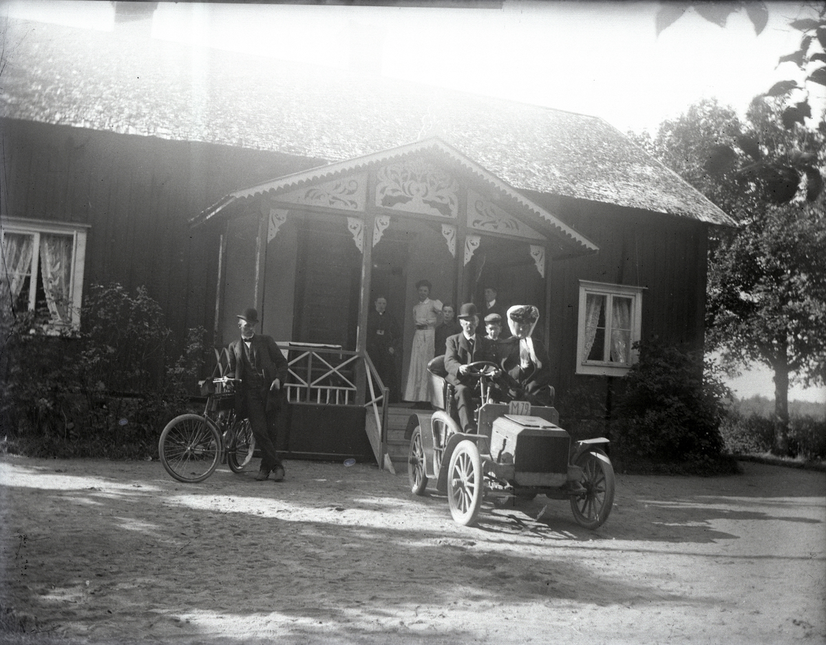 En bil med registreringsnummer M 79 är parkerad framför ett bostadshus på landet, troligen i närheten av Urshult.
Bilen är en Scania från cirka 1903, som ägdes av en Johan Sjögren. Bilen fick senare reg.nr. G 5 och skrotades 1919.
Vid cykeln står Johans son Gustaf Sjögren.