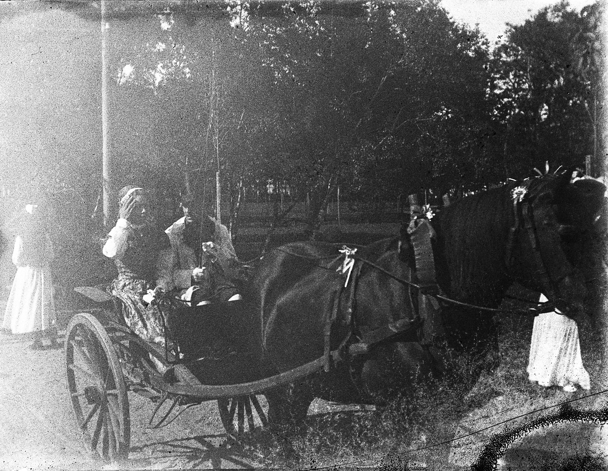 En hästskjuts med två passagerare i folkdräkt. 
Trol. taget i samband med Barnens Dags-firande eller likn. 
Mörkt foto.