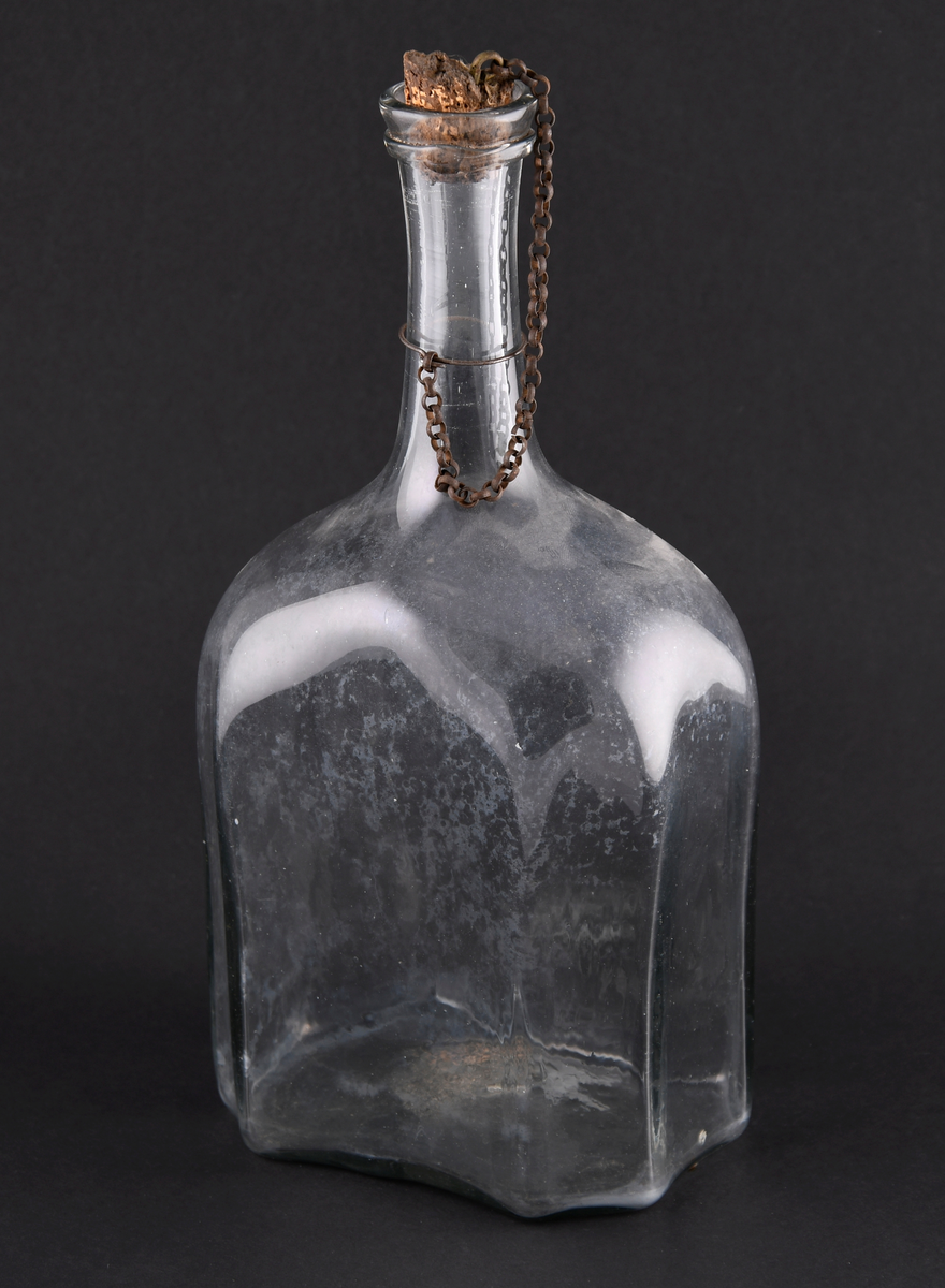Flaske av håndblåst glass. Rektangulær form med avrundede skuldre. Flasken har brutte hjørner, slik at den er åttekantet. Krage under helletuten. Flasken har en kork med kjetting som går rundt halsen av flasken.