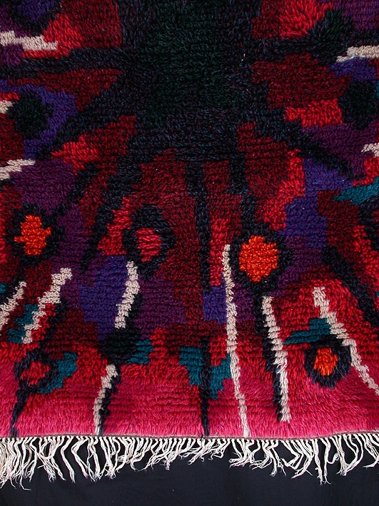Rektangulär ryamatta i cerise, rosa och rött med från den svarta mitten strålande mönster i svart, lila, grönt, beige och orange. 130 mm långa linfransar i kortsidorna av varpen.