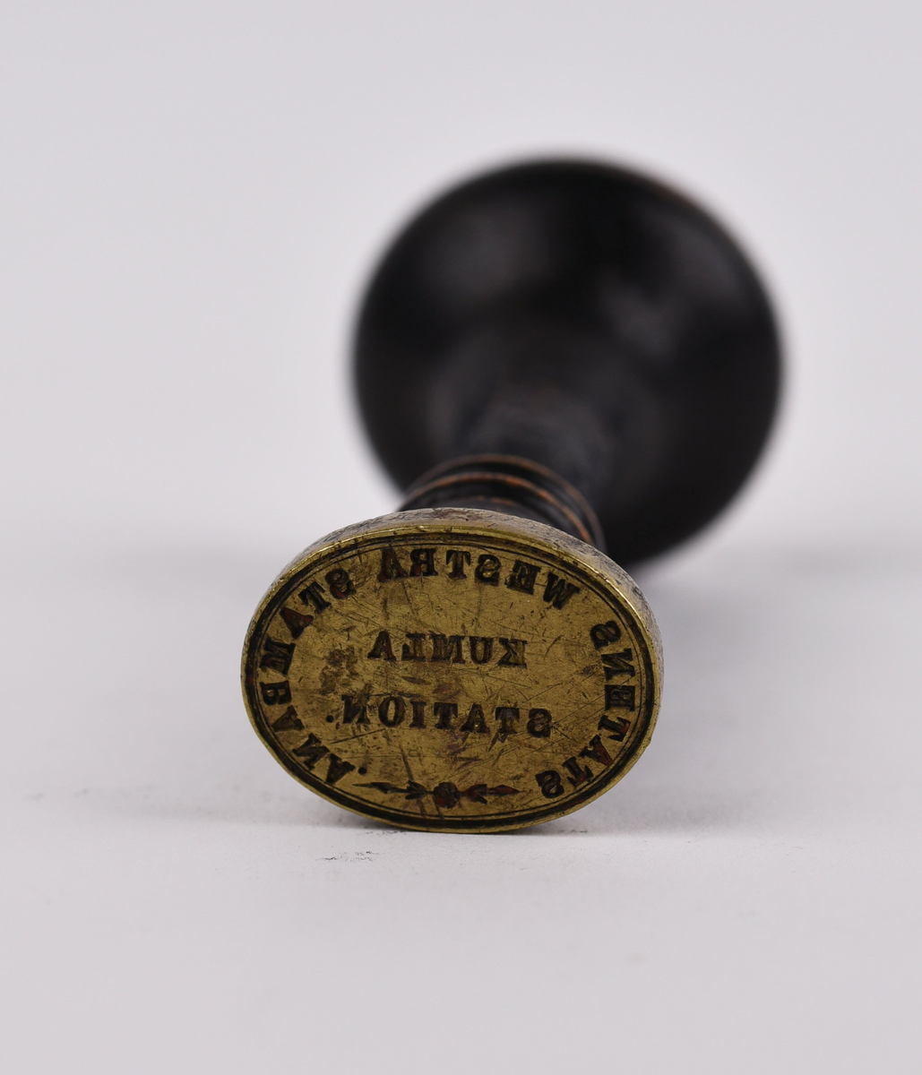 Metallstämpel med svarvat trähandtag. Oval fot av mässing med texten "STATENS WESTRA STAMBANA KUMLA STATION". Följande märkning finns på ovansidan "XII/136".