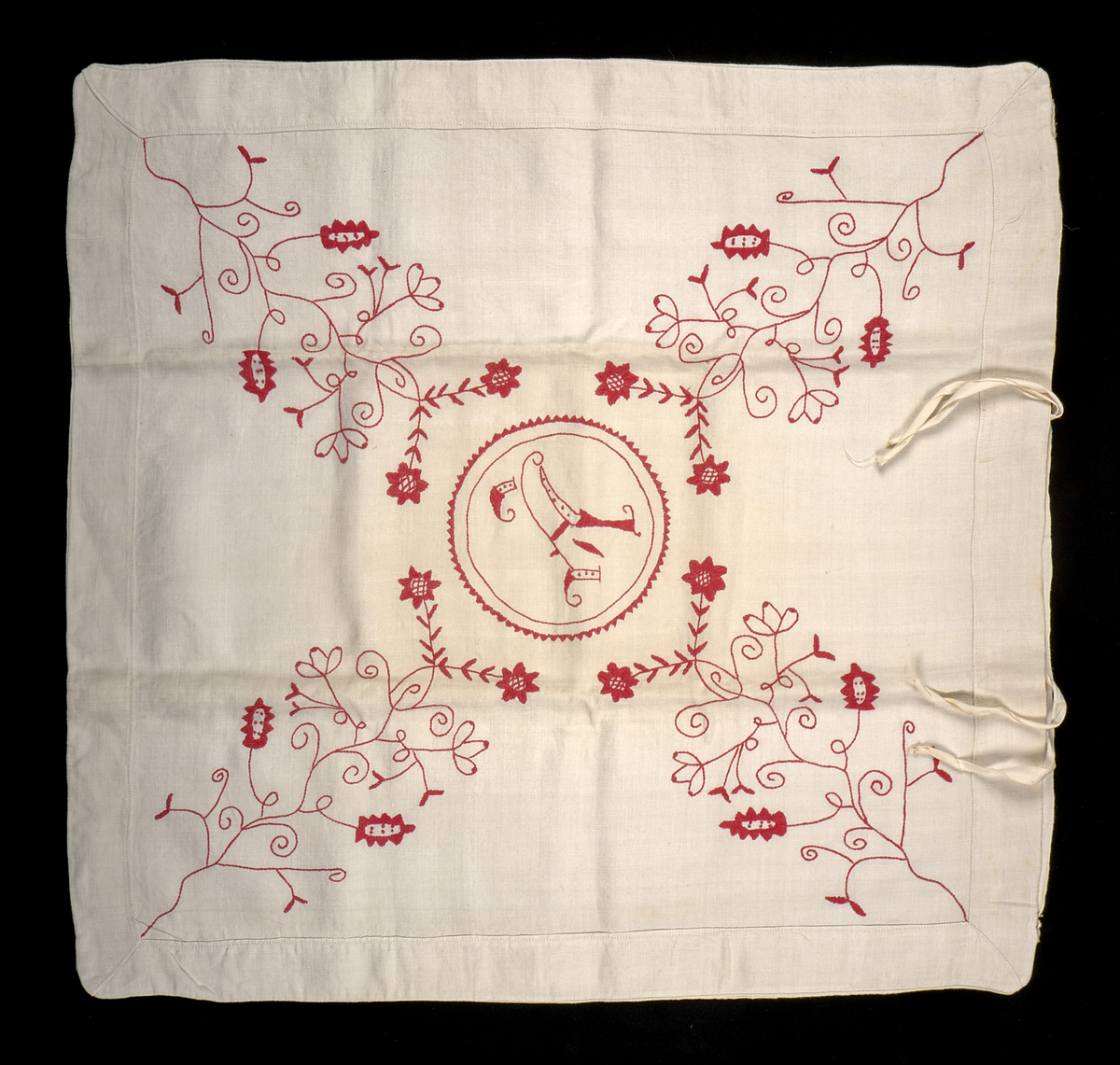 Kuddvar i vit linnelärft, dekorerat på ovansidan med broderi i hallandssöm utfört med rödfärgad bomullstråd. I mitten av motivet en bokstav omsluten av en cirkel: "A". Kuddvaret är sammansytt av två tygstycken, fram- och baksida, och har på båda sidorna vikta fållar längs kanterna.