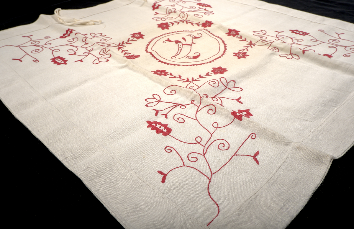 Kuddvar i vit linnelärft, dekorerat på ovansidan med broderi i hallandssöm utfört med rödfärgad bomullstråd. I mitten av motivet en bokstav omsluten av en cirkel: "A". Kuddvaret är sammansytt av två tygstycken, fram- och baksida, och har på båda sidorna vikta fållar längs kanterna.