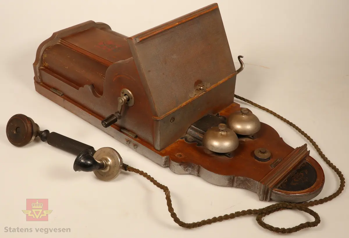 Telefonapparat festet til en treplate som er ment til å henges opp på veggen. Treplaten er brunbeist. Telefonapparatet har to bjeller på. Sort og blankt telefonrør. Sveivehåndtak på siden. Den nedre delen av apparatet er preget med våpenskjold og navn på produsent. 